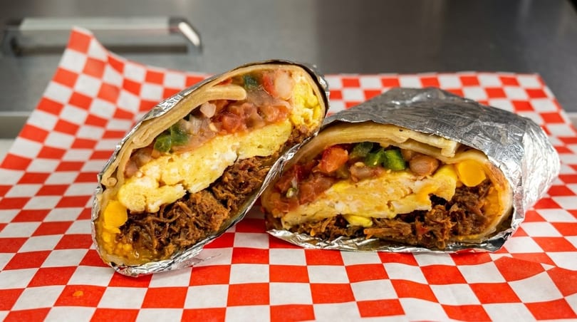 Two halves of a breakfast burrito, filled with eggs, meat, and salsa, on red and white checkered paper.